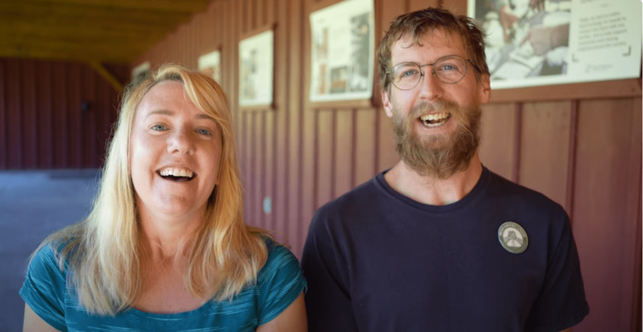 A woman and a man smile in front of a red building