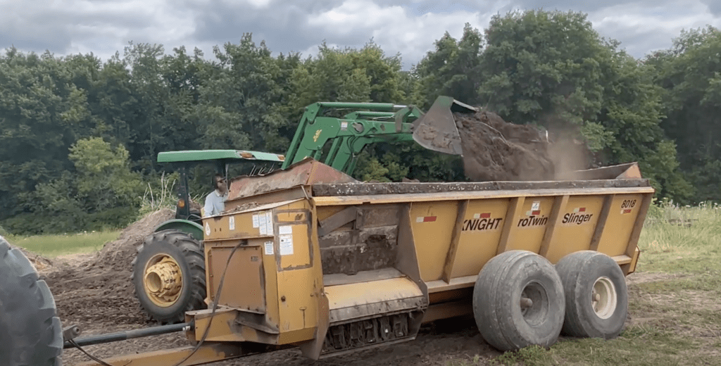 A green tractor dumps compost into a large yellow "Knight" manure spreader trailer