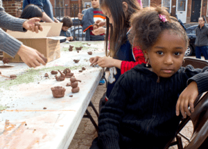 A young girl sits next to a folding table with several small clay structures, with other people working with the clay in the background
