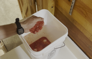 A hand squeezes the seeds and pulp out of a halved tomato into a white bucket