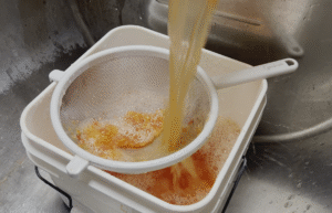 Tomato seeds and pulp being poured through a mesh strainer into another bucket in a sink.
