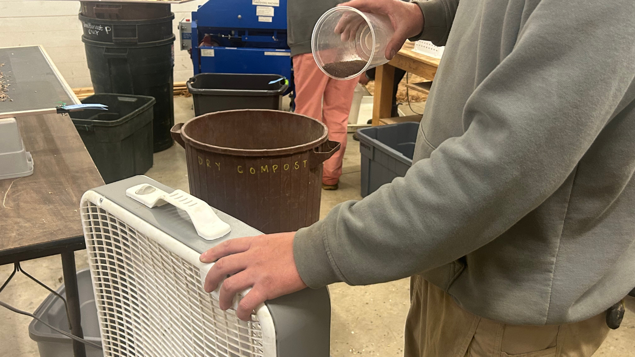 A person drops tiny petunia seeds out of a plastic cup in front of a box fan winnowing set up